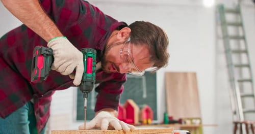 Concentrated Young Bearded Carpenter Wearing Jeans and a Plaid Flannel Shirt Drilling a Hole in a