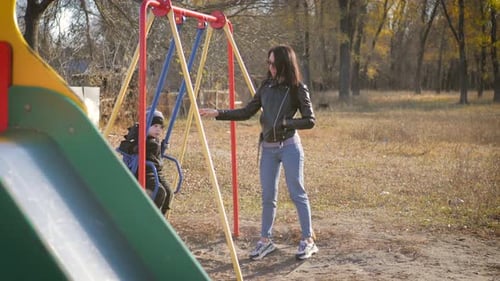 Junge Mutter schüttelt ihren kleinen Sohn auf einer Schaukel in einem Herbstpark. Konzept der glücklichen Kindheit.