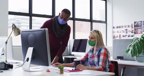 Diverse male and female office colleagues wearing face masks using computer at modern office