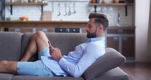 Young Man Holding Smartphone Device Texting Sms Message Lying in Couch at Home Office Smiling Guy