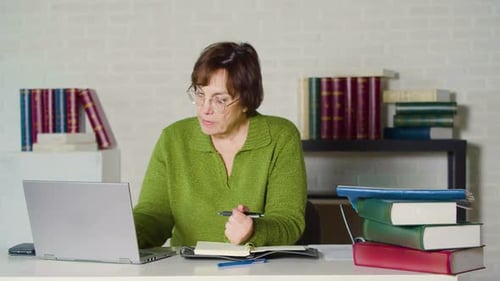 Senor Adult Woman in a Green Sweater Works in Front of a Laptop Monitor in the Evening Office