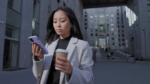 Junge erwachsene asiatische Frau, die auf ihrem Smartphone surft und ihre Augen vor einem Business Center hebt