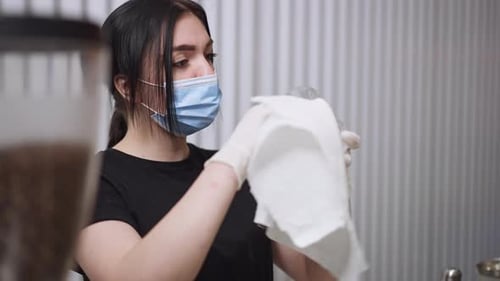 Young Caucasian Slim Woman in Covid Face Mask Rubbing Glass in Cafe at Workplace