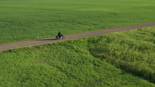 Motorbiker Rides on Ground Road Past Green Fields at Sunset