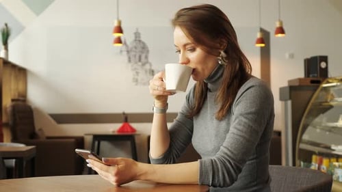 Young Woman in Cafe is Typing a Message on Phone and Drinking a Cup of Coffee Waiting Somebody