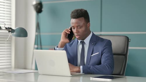 African Businessman Talking on Smartphone While Using Laptop in Office