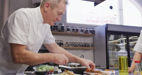 Caucasian male chef wearing chefs whites in a restaurant kitchen, putting food on a plate