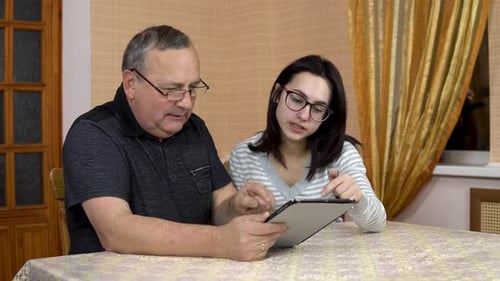 A Daughter Teaches Her Father How To Use a Tablet. A Young Woman Shows Her Old Father Where To Click