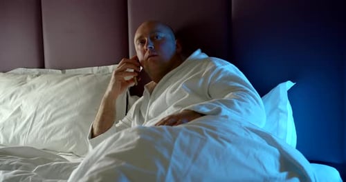 Homme chauve adulte qui s'est récemment réveillé, parlant sur un téléphone portable, allongé sur le lit, tenant un