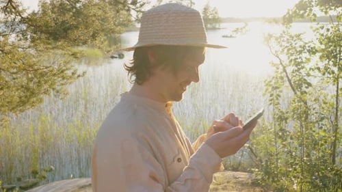 Man in Hat Tries to Get Cell Signal on Phone on River Bank