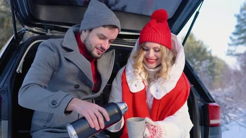 Young Couple a Man and a Woman are Sitting in the Trunk of a Car in a Winter Snowy Forest Hugging