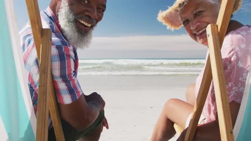 Mixed race senior couple sitting on sunbeds and smiling at the beach