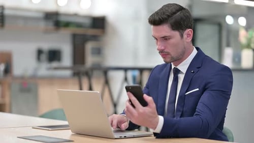 Businessman Using Smartphone and Laptop in Office