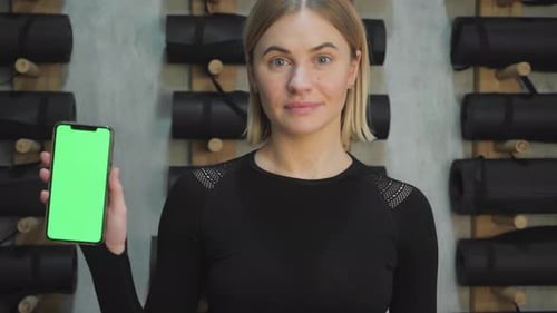 Happy young beautiful blonde woman showing a smartphone with a green screen in the gym