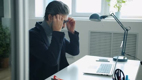 Mature Man in Elegant Suit Putting on Glasses and Working with Laptop in Office