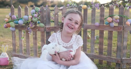 Nahaufnahme eines Mädchens in einem Osterkleid, das lächelt und einen Osterhasen vor einem Lattenzaun hält