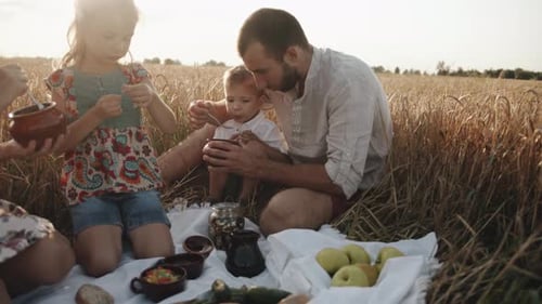 A Young Happy Family with Children is Relaxing on a Picnic in a Wheat Field and Eating the Food They