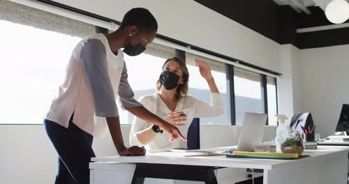 Two diverse female colleagues wearing face masks looking at laptop and discussing in office