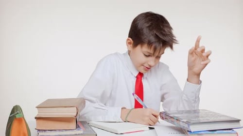 Young Caucasian Kid School Boy in Red Tie Sitting at Table Learning Writing Surrounded By Books on