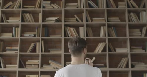 Jeune homme brun debout devant des étagères dans la bibliothèque et choisissant la littérature. Déplacements de caméra