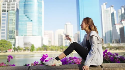 4K Asian woman drinking water from a bottle while jogging at public park in the morning.