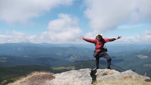 Tourist with Backpack on the Top of Mountain Near the Cliff Raises Hands To Side