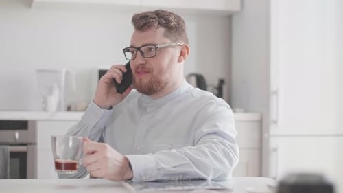 Joven elegante vestido con ropa ligera se comunica por teléfono para desayunar en la cocina de su casa