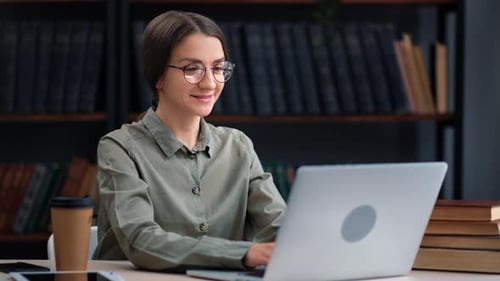 Lächelnde selbstbewusste Wirtschaftsstudentin mit Brille beim Chatten Verwenden Sie Laptop am Schreibtisch der Bibliothek