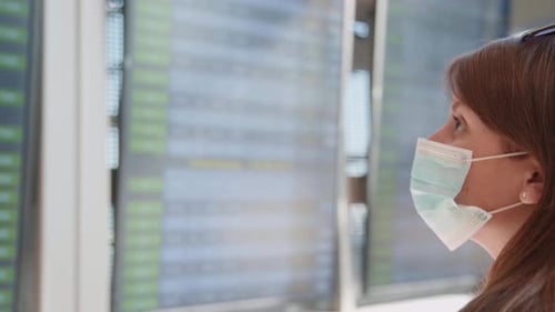 Young Woman Looks at Airport Flight Board Wearing Mask