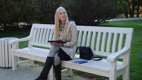 Young Woman Sits On The Bench In Autumn Park And Works On The Tablet