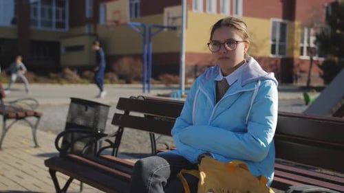 A Sad Teenage Girl is Sitting on a Bench in the School Yard Against the Background of Peers Playing