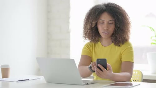 African Woman Browsing Internet on Smartphone while using Laptop in Office