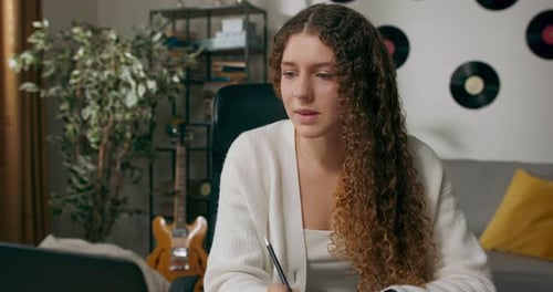 Girl with Curly Hair Sits at Desk in Living Room and Works on Laptop Taking Notes Focused Young