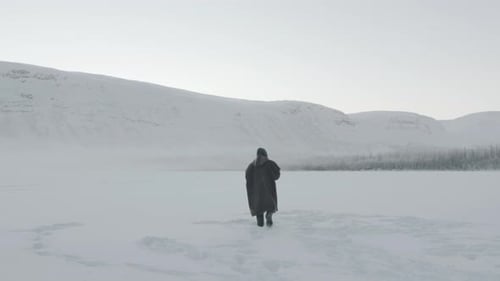 A Man in a Wide Coat with a Hood Goes Along a Snowy Field Against the Backdrop of a Mountain