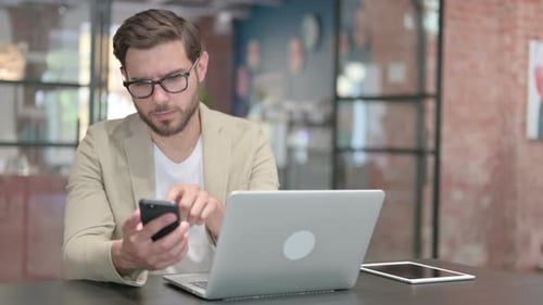 Man Using Phone at Desk with Laptop
