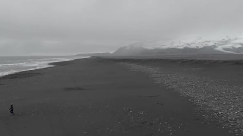 Aerial shot of a woman running along a black sand beach with panning towards the mountains.