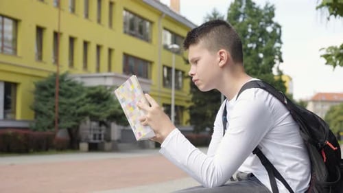 A Caucasian Teenage Boy Reads a Book Side View a School in the Background