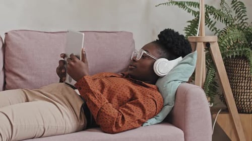 Frau mit Kopfhörern liegt auf Sofa und hört Musik auf dem Tablet