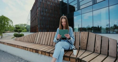Woman is Sitting on a Park Bench and Scrolling on the Tablet in Her Hands