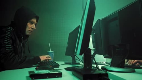 Two Hackers in Hoods Work on a Computers with Maps and Data on Display Screens in a Dark Office Room