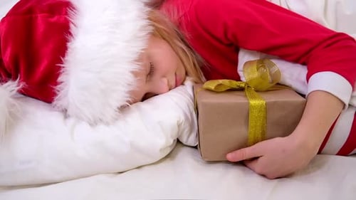 Menina de chapéu de Papai Noel e pijama de Natal deitada na cama com caixa de presente em casa
