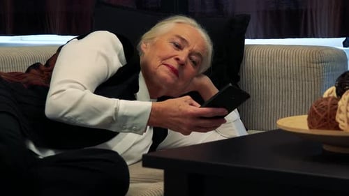 Old Caucasian Woman Lies on Couch and Works on Smartphone in Living Room