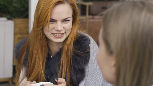 Two Cute Caucasian Girlfriends Covered in Blankets Sitting in the Cafe Chatting and Drinking Hot Tea