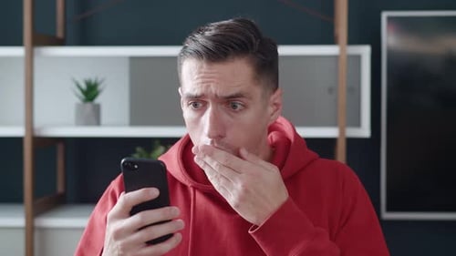 Frightened Hipster Man Reacting Message on His Smartphone Covers His Mouth in Shock at Home Office