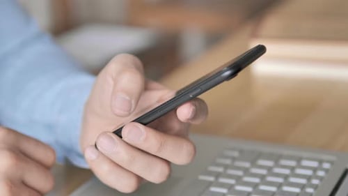 Hands Using Smartphone Next to Laptop at Desk