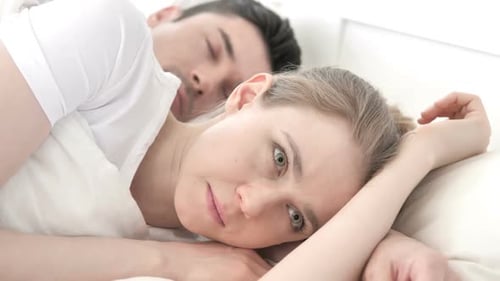Tense Woman Lying in Bed Along Sleeping Boyfriend