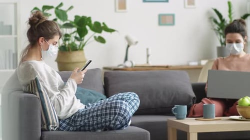 Women in Medical Masks Using Smartphone and Laptop while Sitting on Couch