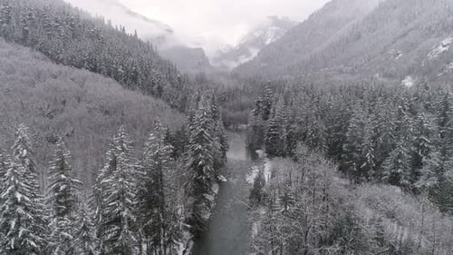 Aerial Of Frozen Winter Landscape In Mountain Forest Valley With Snow Flakes Falling On Cold River