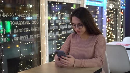 A Young Woman is Sitting at a Table in a Cafe in the Evening and Texting on the Phone