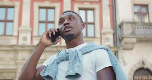 Afroamericano de pie cerca de un antiguo edificio urbano y hablando por teléfono móvil